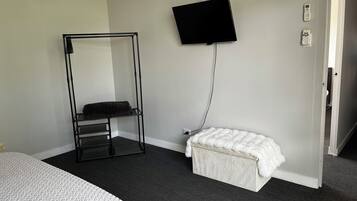 Ein Schlafzimmer mit einem Bett, einem schwarzen Metallregal, einer weißen, flauschigen Bank, einem an der Wand montierten Fernseher und einer Tür zu einem anderen Raum.
