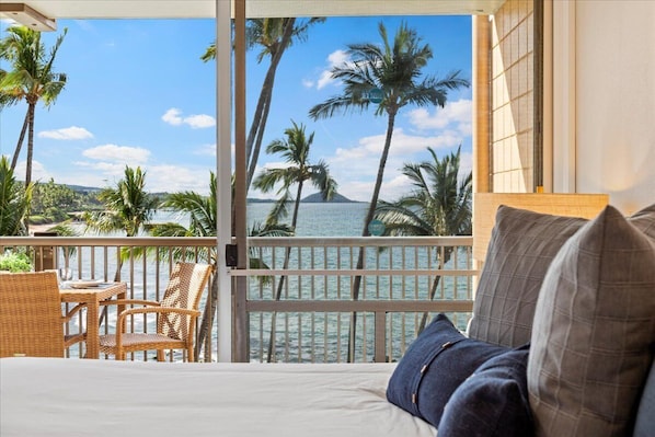 A balcony with a view of the ocean and palm trees, a bed with pillows, and wicker furniture.