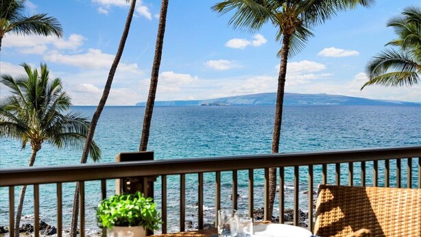 Un balcón con vista al océano, palmeras y una zona de comedor con una planta y vasos.