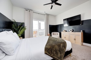 A modern hotel room with a large bed, a ceiling fan, a flat-screen TV, a small side table, a chair, and a view of the city through a large window.