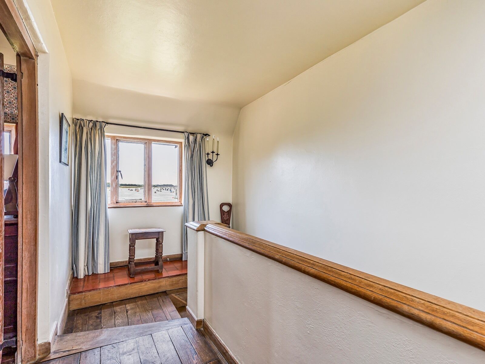 A narrow hallway with a wooden floor, a window with curtains, a small wooden bench, and a wall-mounted light fixture.