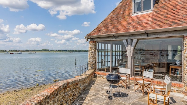 Un bâtiment en pierre, surmonté d’un toit en tuiles, qui surplombe un plan d’eau paisible. Il est doté d’une terrasse avec des meubles d’extérieur et d’un barbecue.