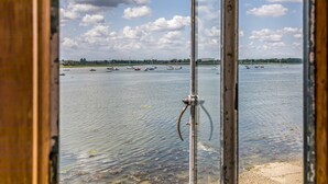 Une vue à travers une fenêtre sur une étendue d’eau où l’on aperçoit des bateaux, un ciel dégagé et une côte lointaine.