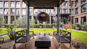 A modern outdoor seating area with swings and a fire pit, surrounded by a well-maintained garden and a building with large windows in the background.