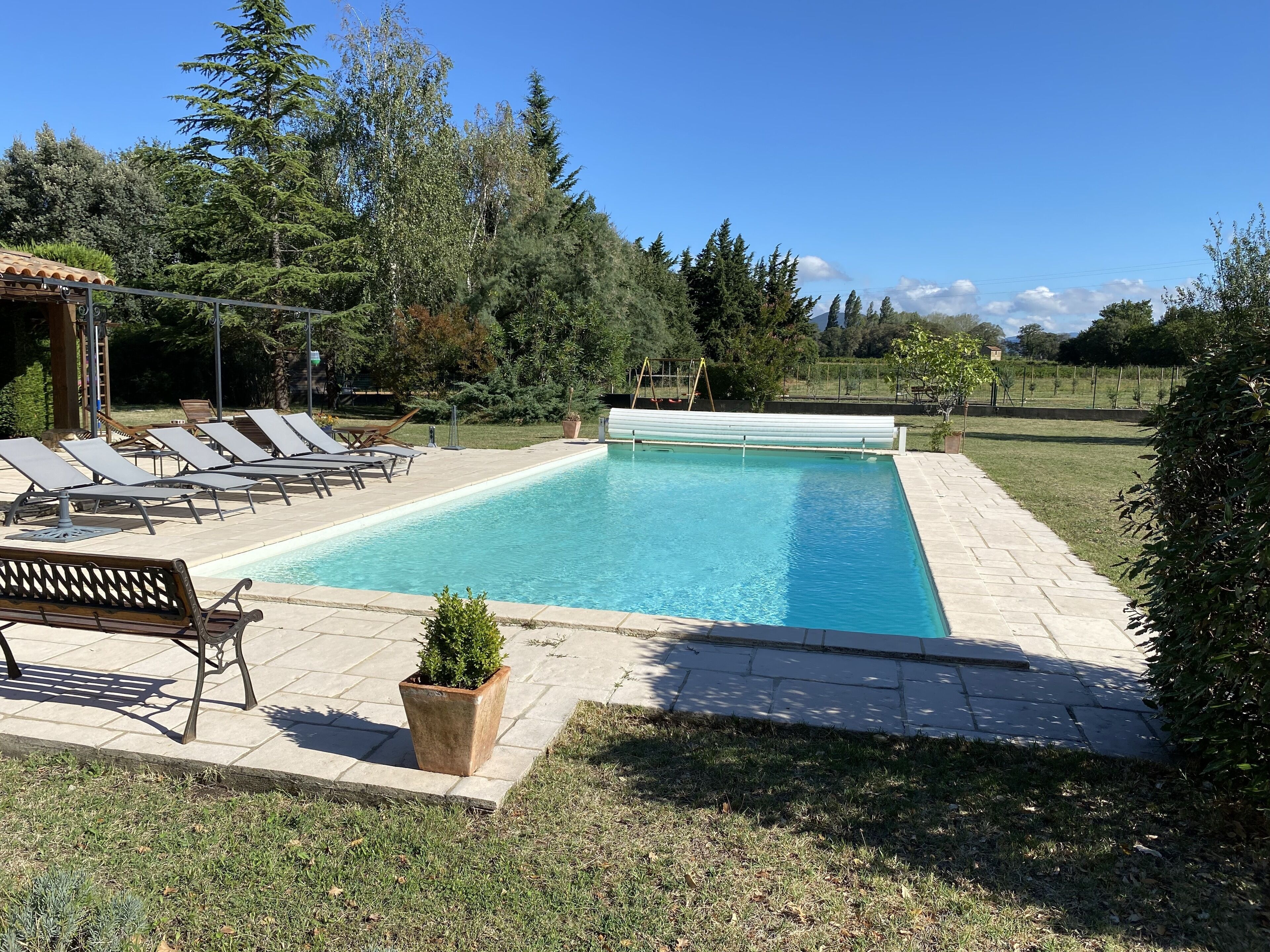 A rectangular swimming pool with a tiled deck, surrounded by lounge chairs and a bench.