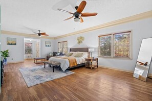 Une chambre spacieuse dotée d’un plancher en bois, d’un grand lit, d’un ventilateur de plafond et d’une fenêtre donnant sur les arbres.