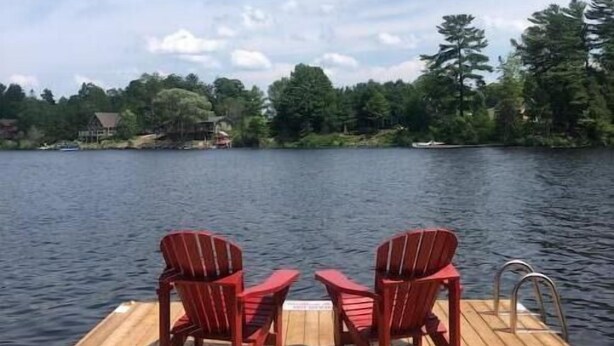 A wooden dock with two red Adirondack chairs facing a calm lake, surrounded by trees and a clear sky. - Lakeside in Muskoka (Huntsville)