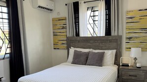 Een net opgemaakt bed met een witte deken, een houten hoofdbord en twee kussens. Er staat een nachtkastje met een lamp en een digitale klok.