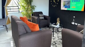 Een moderne woonkamer met een bank, fauteuils, een salontafel en een grote wandklok. Er staat een potplant en een raam met gordijnen.