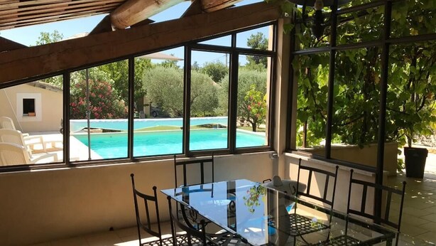 Un tavolo in vetro con sedie in una stanza luminosa, con ampie finestre vista su una piscina e vegetazione lussureggiante.