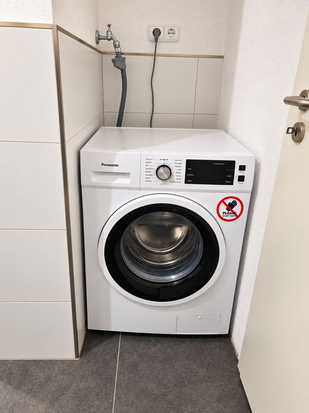 Lavadora blanca compacta con panel de control, una manguera y un cartel que dice 'NO TOCAR'.