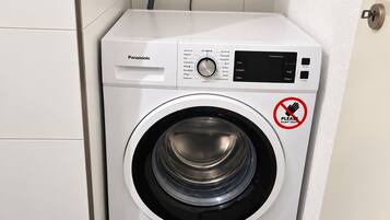 Eine kompakte, weiße Waschmaschine mit Bedienfeld, Schlauch und einem Warnschild mit der Aufschrift 'DO NOT TOUCH'.
