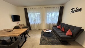A modern living room with a wooden dining table, a grey sofa with red cushions, a coffee table, and a TV mounted on the wall.