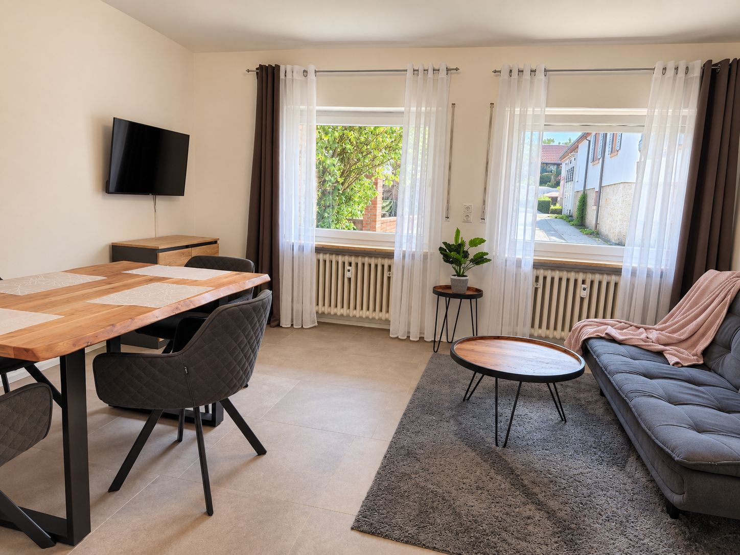 Een moderne woonkamer met een eethoek, een bank, een aan de muur bevestigde televisie en uitzicht op het buiten.