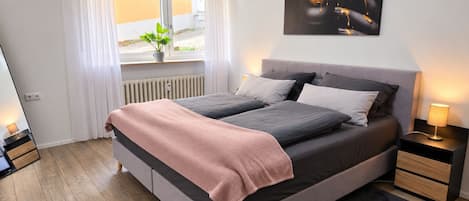 A modern bedroom with a large bed, a bedside table, a nightstand, a window with curtains, and a wall-mounted artwork.