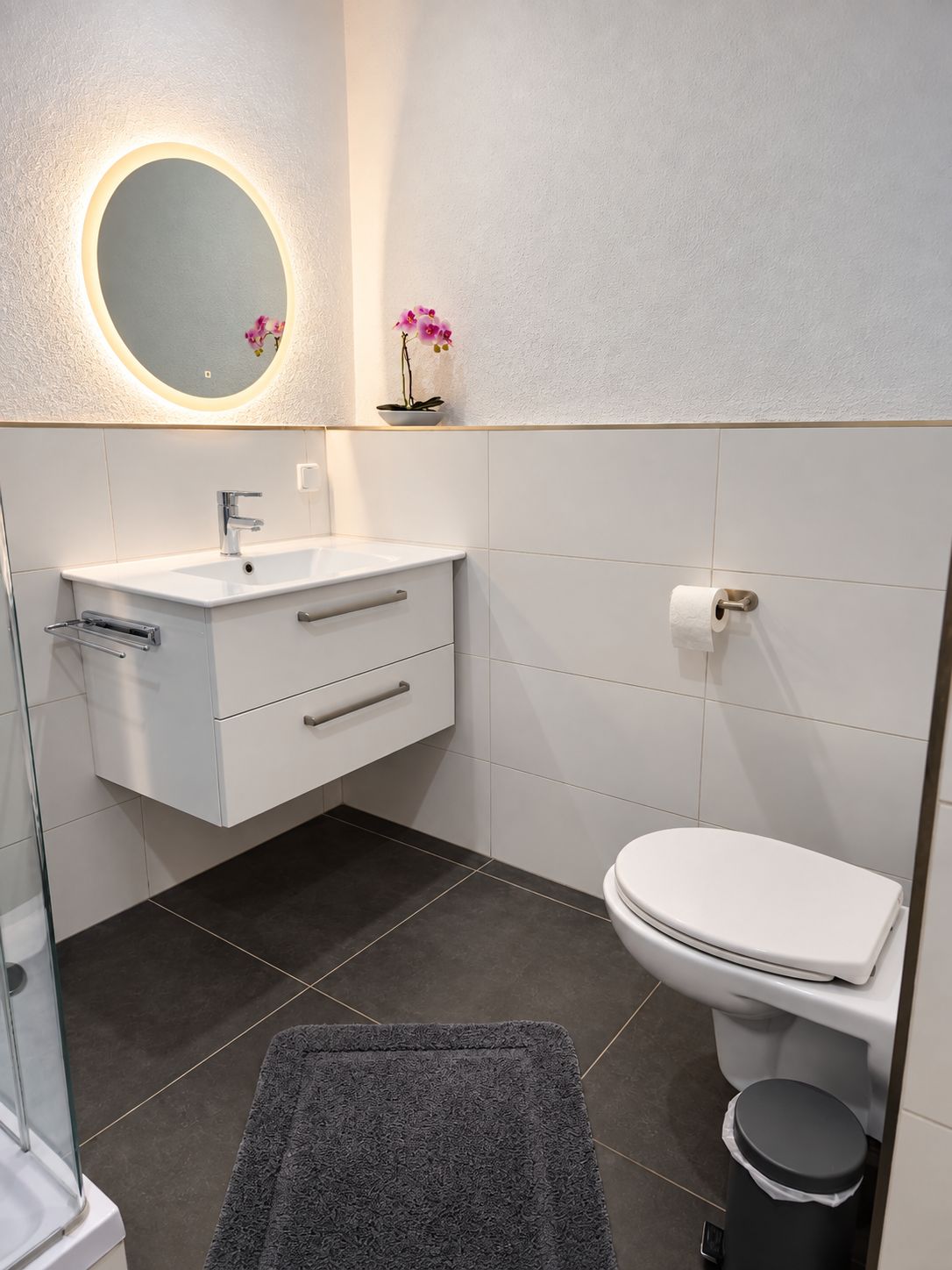 Un bagno moderno con un mobile lavabo bianco, uno specchio rotondo illuminato, un water e un cestino.
