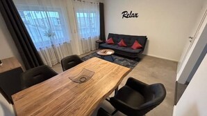 A modern living room with a wooden dining table, black chairs, a grey sofa with red cushions, and a 'Relax' sign on the wall.