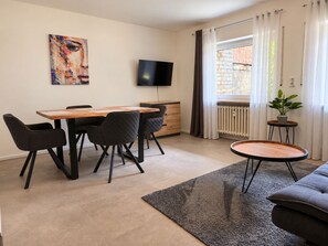 A modern living room with a dining area, a flat-screen TV, a potted plant, and a view of the outside.