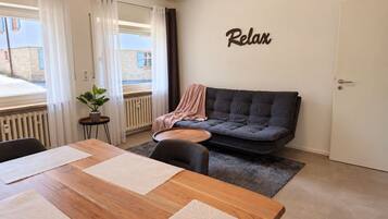 Ein modernes Wohnzimmer mit einer Couch, einem Esstisch mit Unterlagen, einer Topfpflanze und einem Wandschild mit der Aufschrift 'Relax'.