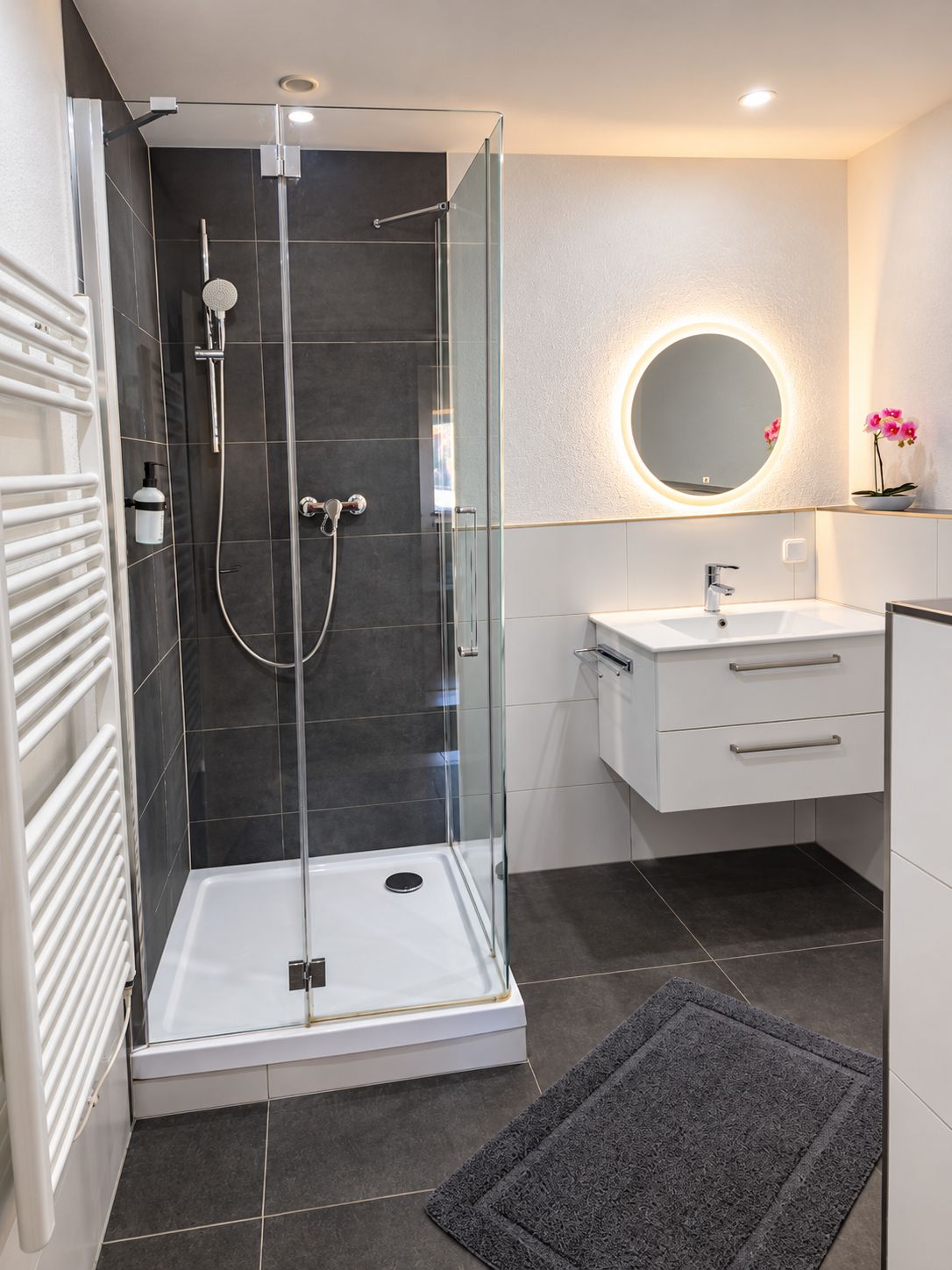 A modern bathroom with a glass-enclosed shower, a white vanity with a round mirror, and a dark tiled floor.