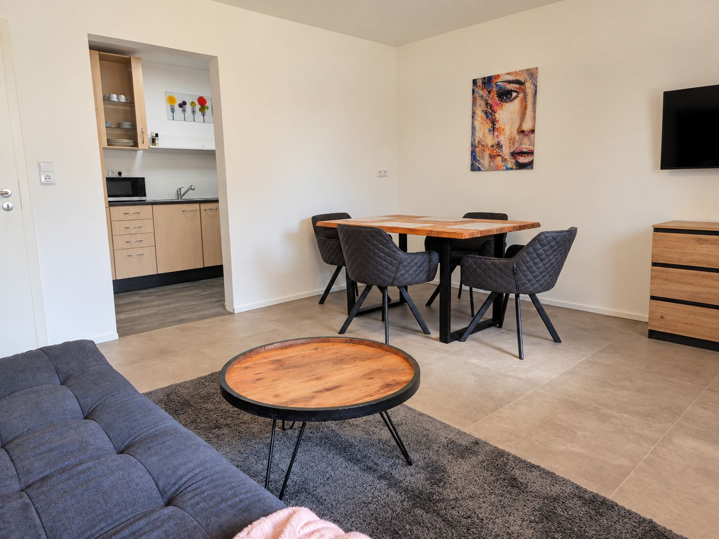 Ein moderner Wohnraum mit einer Couch, einem runden Couchtisch, einem Esstisch mit Stühlen, einer Küchenzeile und einem Bild an der Wand.