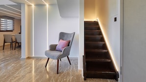 Un intérieur moderne avec un sol en marbre, un fauteuil recouvert d’un tissu gris agrémenté d’un coussin rose, et un escalier dont les bords sont illuminés.