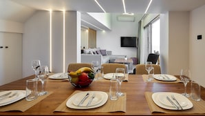 Un coin repas avec une longue table en bois, dressée pour quatre personnes, entourée de chaises, et agrémentée d’un saladier de fruits et de verres à vin posés sur la table.