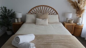 Cama bem arrumada com colcha bege, toalha branca enrolada e duas mesas de cabeceira com abajures e um pequeno vaso com plume de penas.