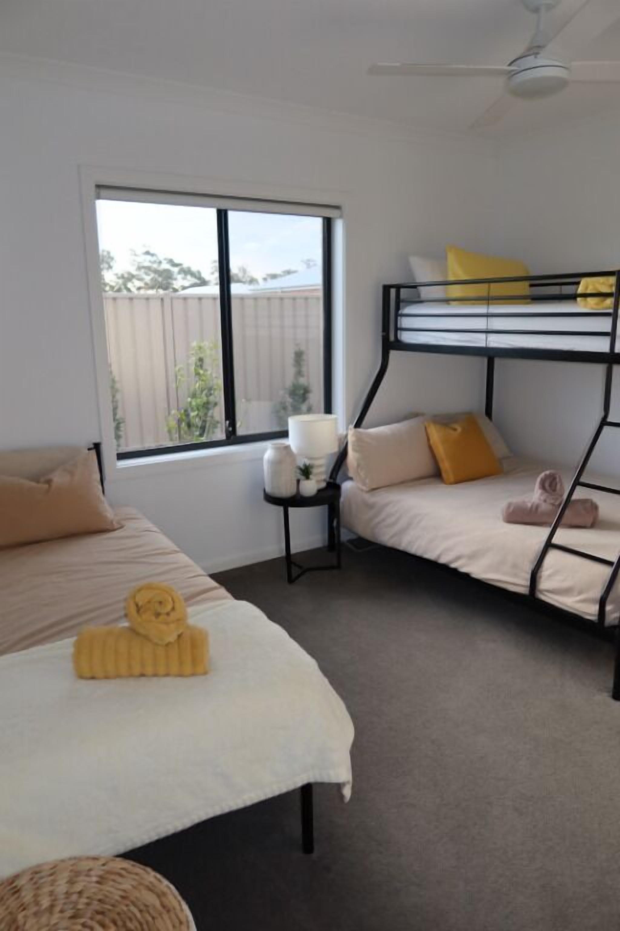 Une chambre avec un lit superposé, un lit simple, une fenêtre avec des stores, un ventilateur de plafond et une petite table avec une lampe.