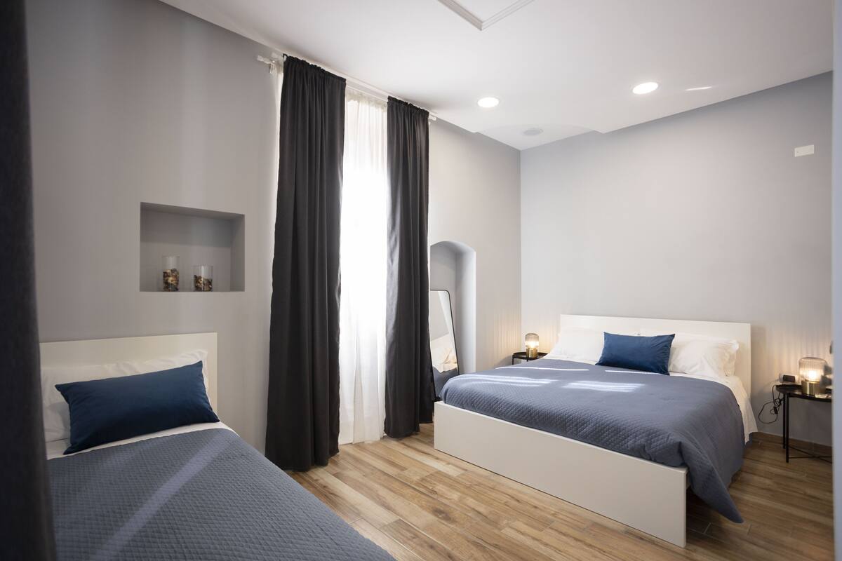 A modern hotel room with two beds, a bedside table with a lamp, a small shelf with decorative items, and a large mirror on the wall.