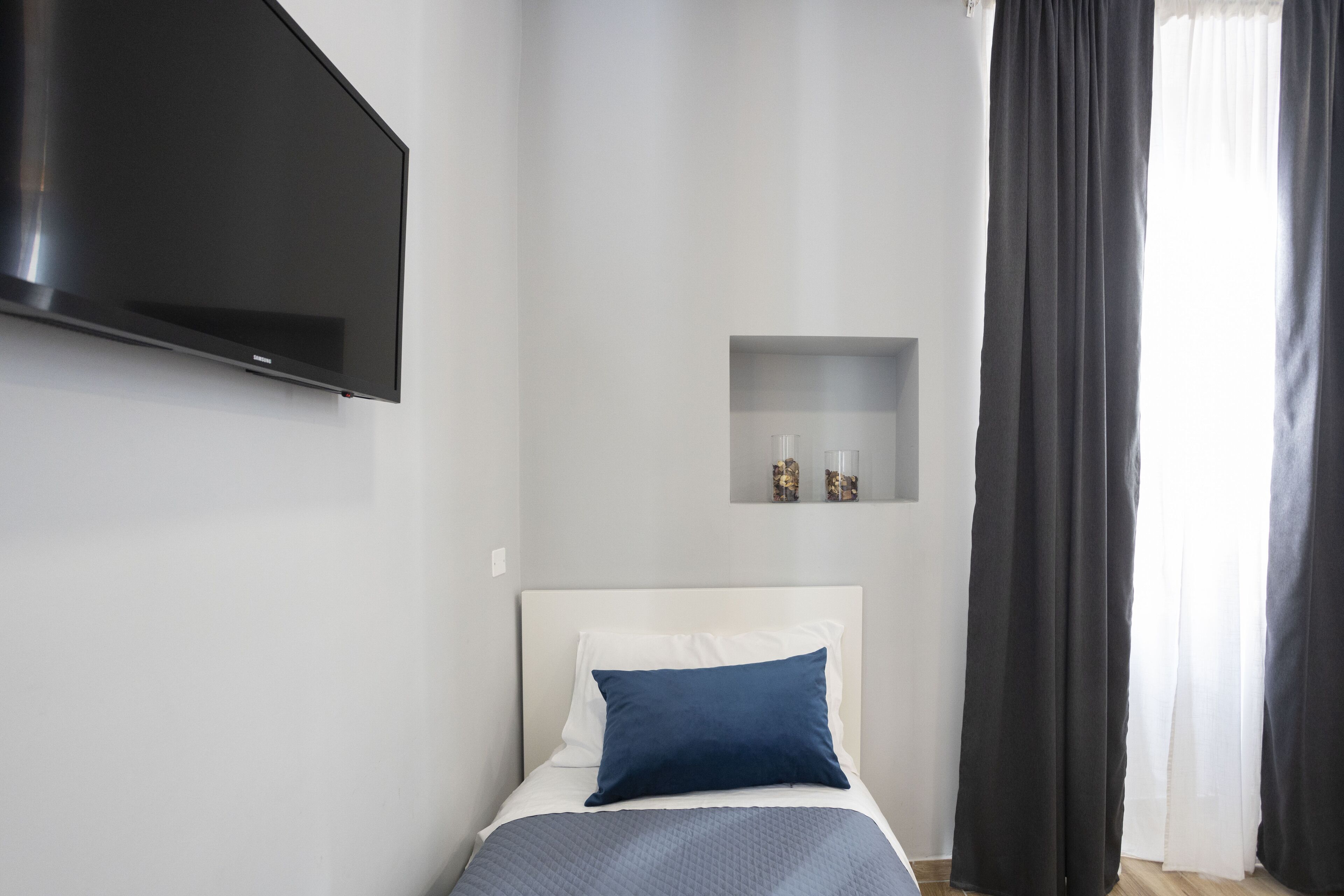 A modern bedroom with a bed, a flat-screen TV mounted on the wall, a small shelf with decorative items, and a window with curtains.