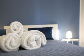 Un lit bien fait, avec des serviettes blanches pliées en rouleaux, et un mur bleu foncé en arrière-plan.