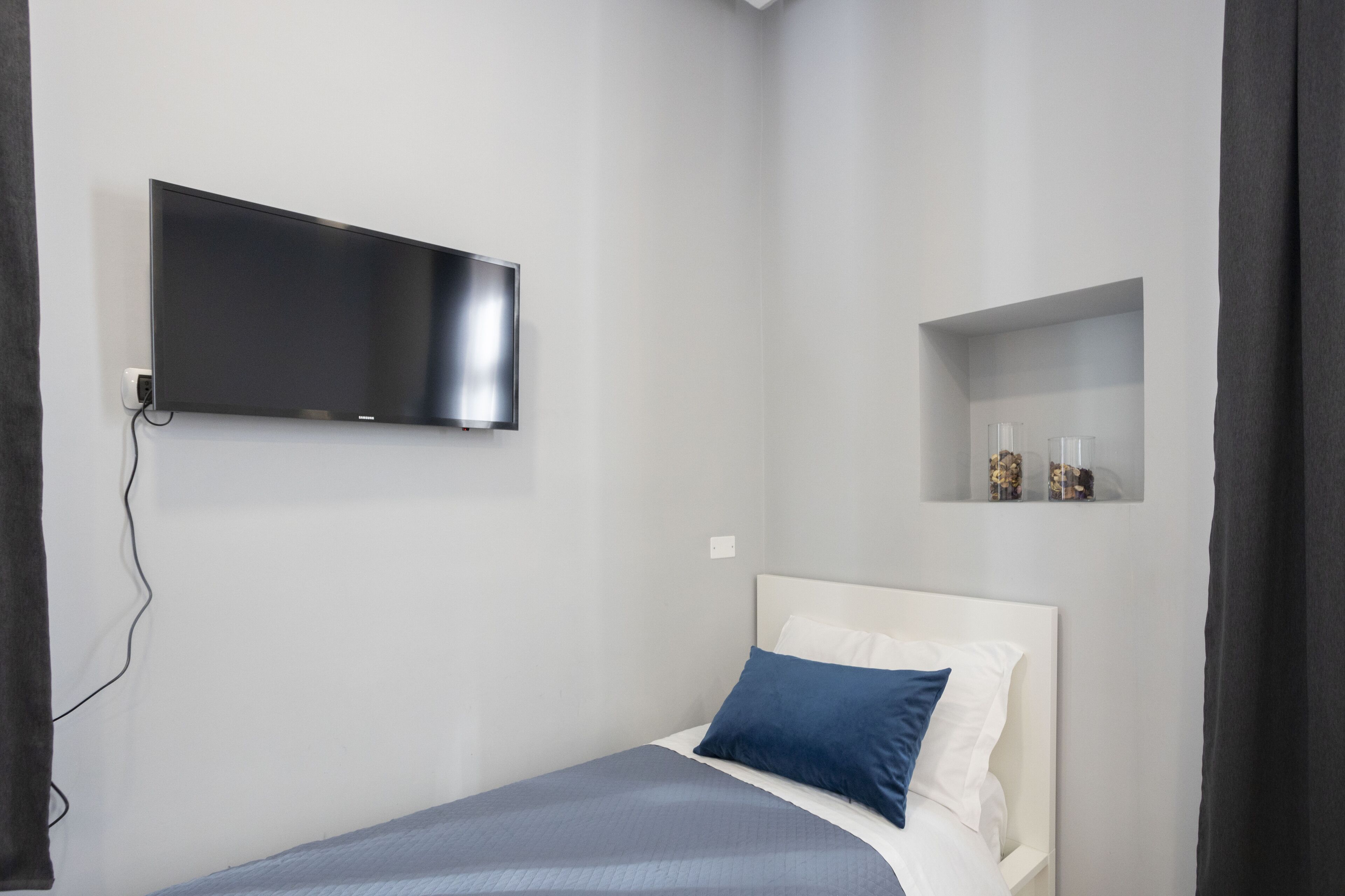 Une chambre à coucher avec un lit, un téléviseur fixé au mur et une petite étagère ornée d’objets décoratifs.