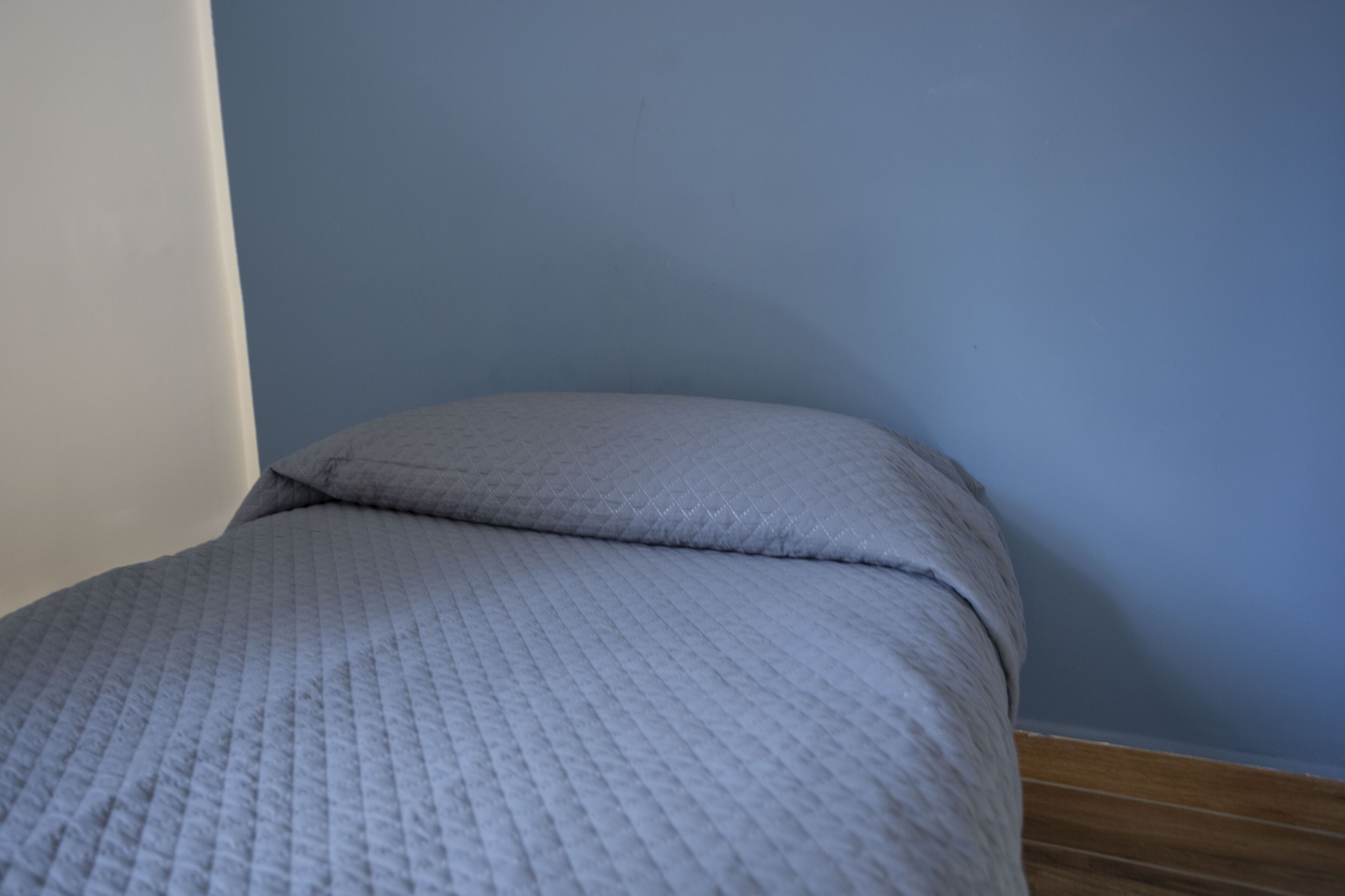 Una cama con cubrecolcha acolchada y almohada frente a una pared azul y piso de madera.