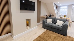 Una sala moderna con un televisor de pantalla plana montado en la pared, un sofá gris con cojines blancos, piso de madera y una escalera que sube a otro nivel.