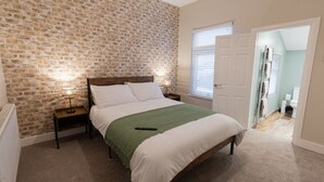 Un dormitorio con pared de ladrillo, cama con ropa de cama blanca, manta verde, dos lámparas de noche y una puerta que da a un baño.