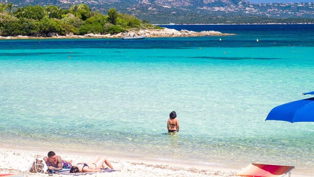 En strand med klart turkis vand, en person stående i vandet og bjerge i baggrunden.