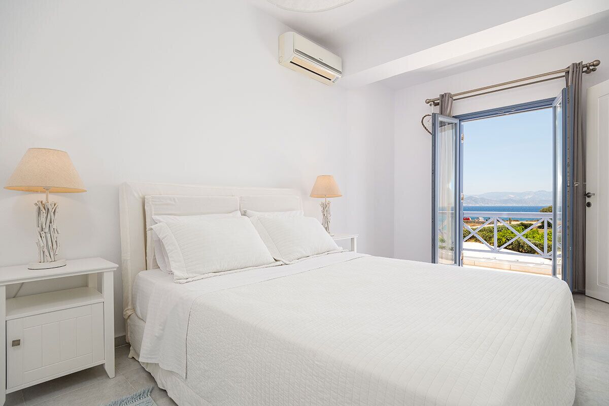 A white bedroom with a bed, nightstands, and an open door leading to a balcony with a view.