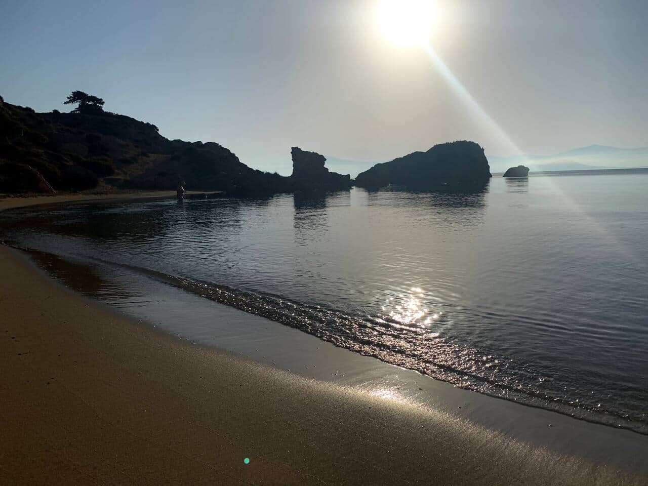 Une plage avec une eau calme, un ciel dégagé et des formations rocheuses qui se dessinent à l’horizon.