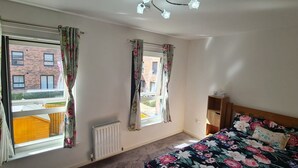 A bedroom with two windows, a radiator, a wooden bed frame, and a bedside table. - Pennywell 2 Bedroom House - Edinburgh  (Edinburgh)