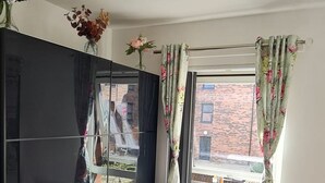 A room with a large black wardrobe, a window with floral curtains, and a view of a brick building outside. - Pennywell 2 Bedroom House - Edinburgh  (Edinburgh)