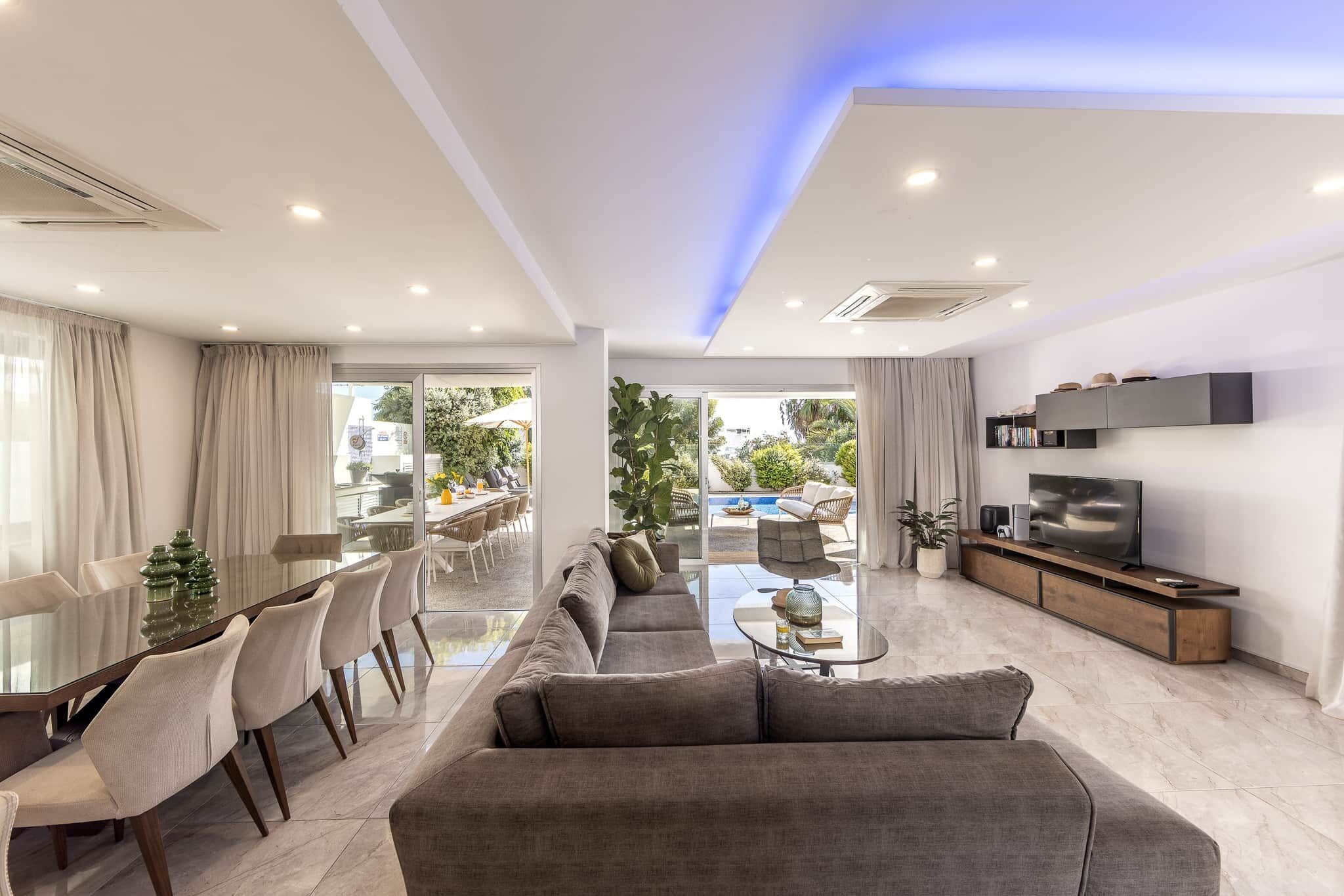 A modern living room with a sofa, dining table, and chairs, a flat-screen TV, and a view of the outdoors.