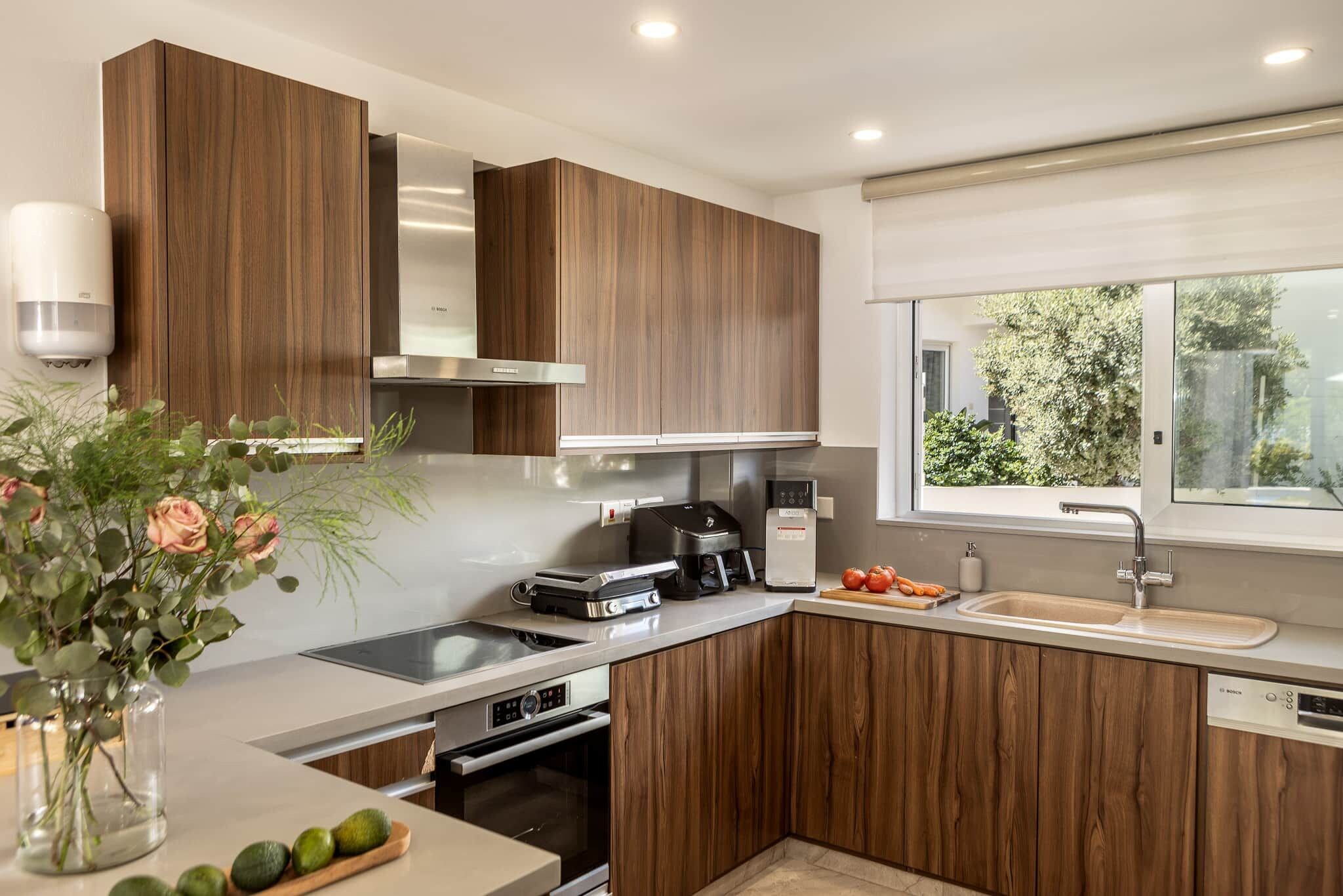 A modern kitchen with wooden cabinets, a built-in oven, a coffee machine, and a window with a view of greenery.