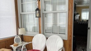 Una habitación acogedora con dos sillas blancas y cojines rojos, una mesita blanca con una lámpara y una ventana que ofrece vistas a una escalera.