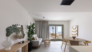 A modern living room with a sofa, a coffee table, and a dining table. There is a large window with curtains, a potted plant, and a wall-mounted artwork. - Morfes Luxury Suites by Caldera Houses (Santorini)