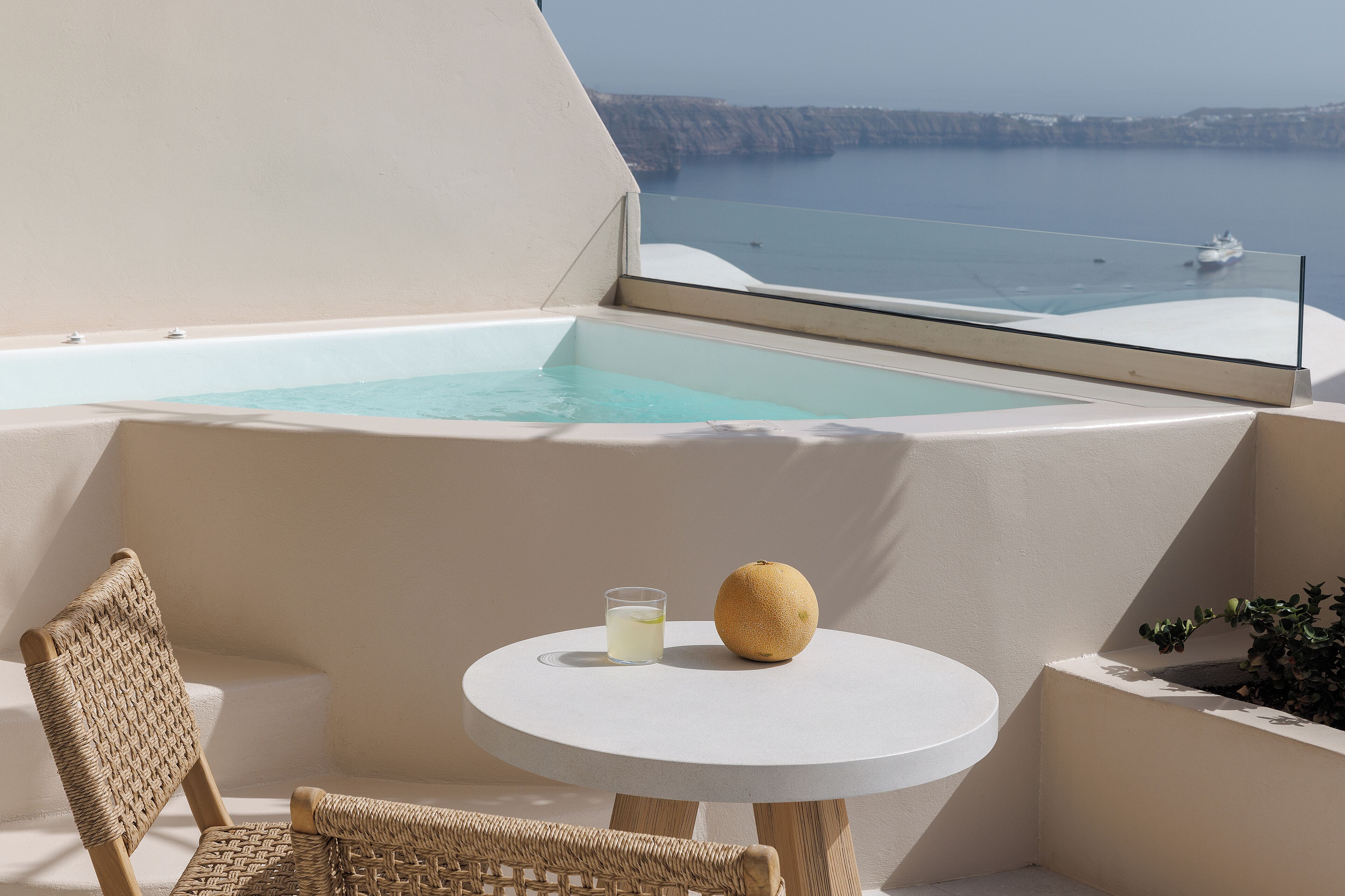 A balcony with a glass-enclosed infinity pool, a wicker chair, a small round table with a glass of water and a fruit, and a view of the sea and cliffs.