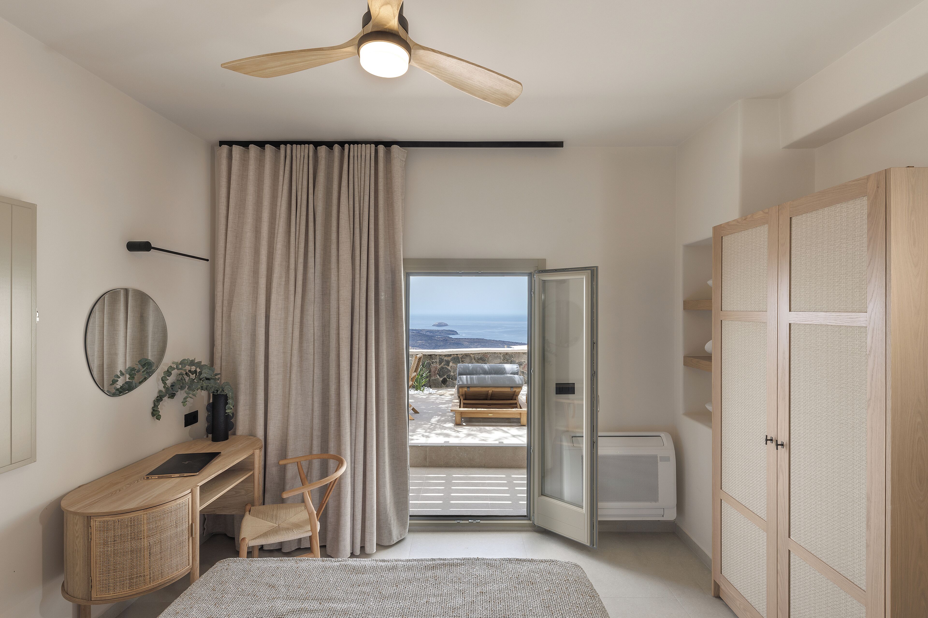 A bedroom with a bed, a desk, a chair, a ceiling fan, a window with curtains, and a view of the outdoors.