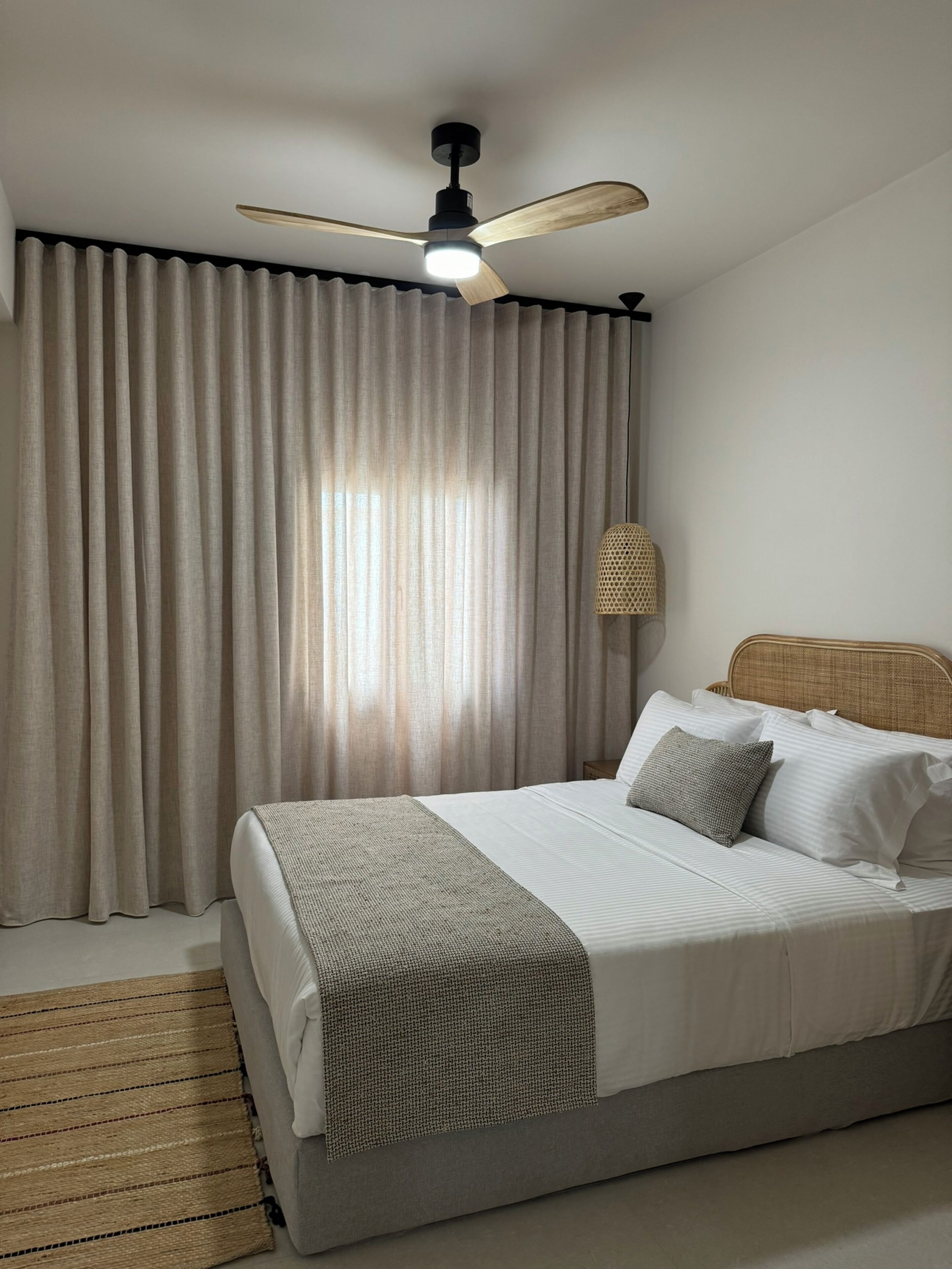 Ein ordentlich bezogenes Bett mit Kopfteil, einem Deckenventilator und einem großen Fenster mit Vorhängen.