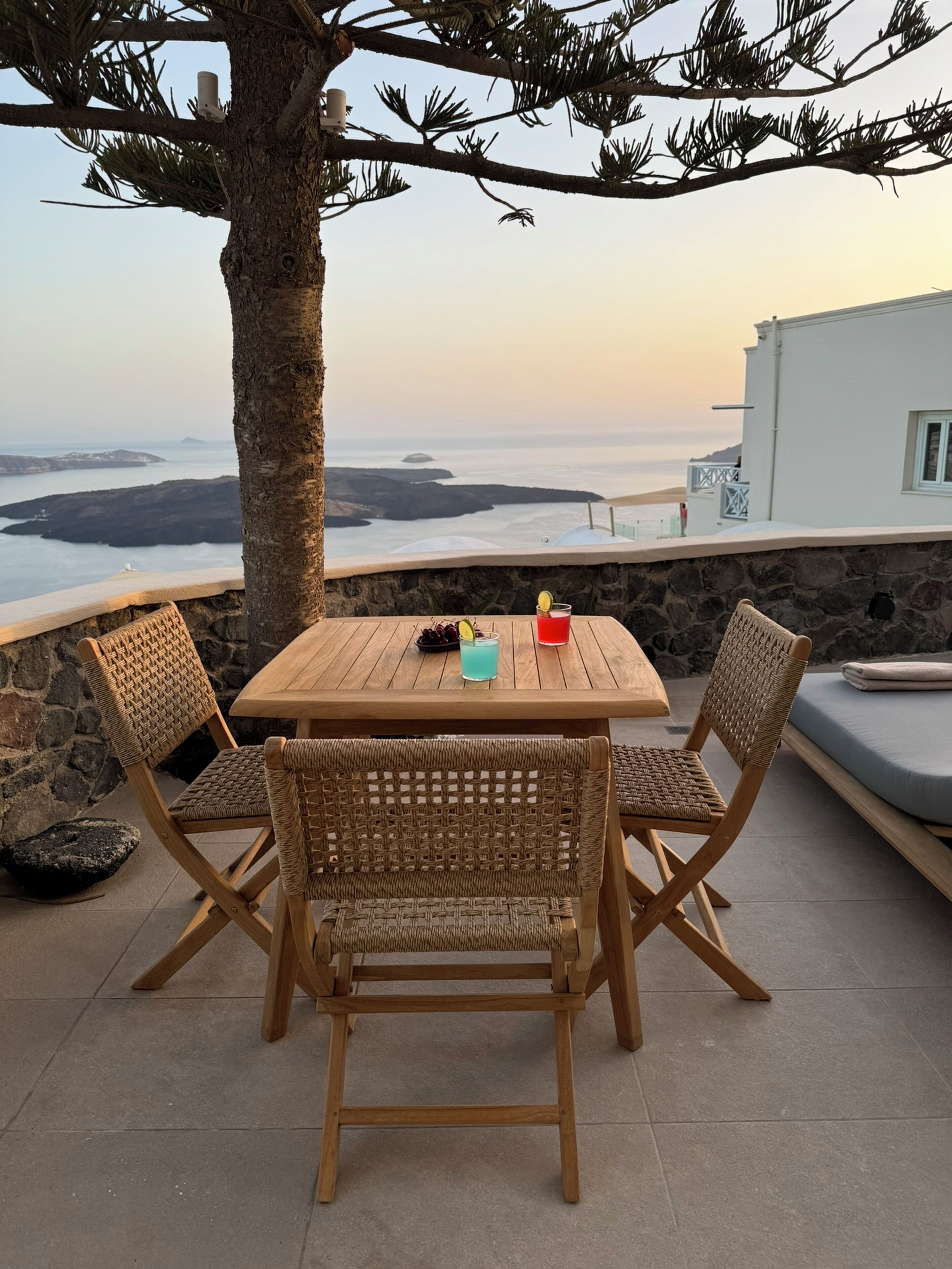 A wooden table with wicker chairs, a drink, and a plate of fruit, overlooking a coastal view with a stone wall and a white building.