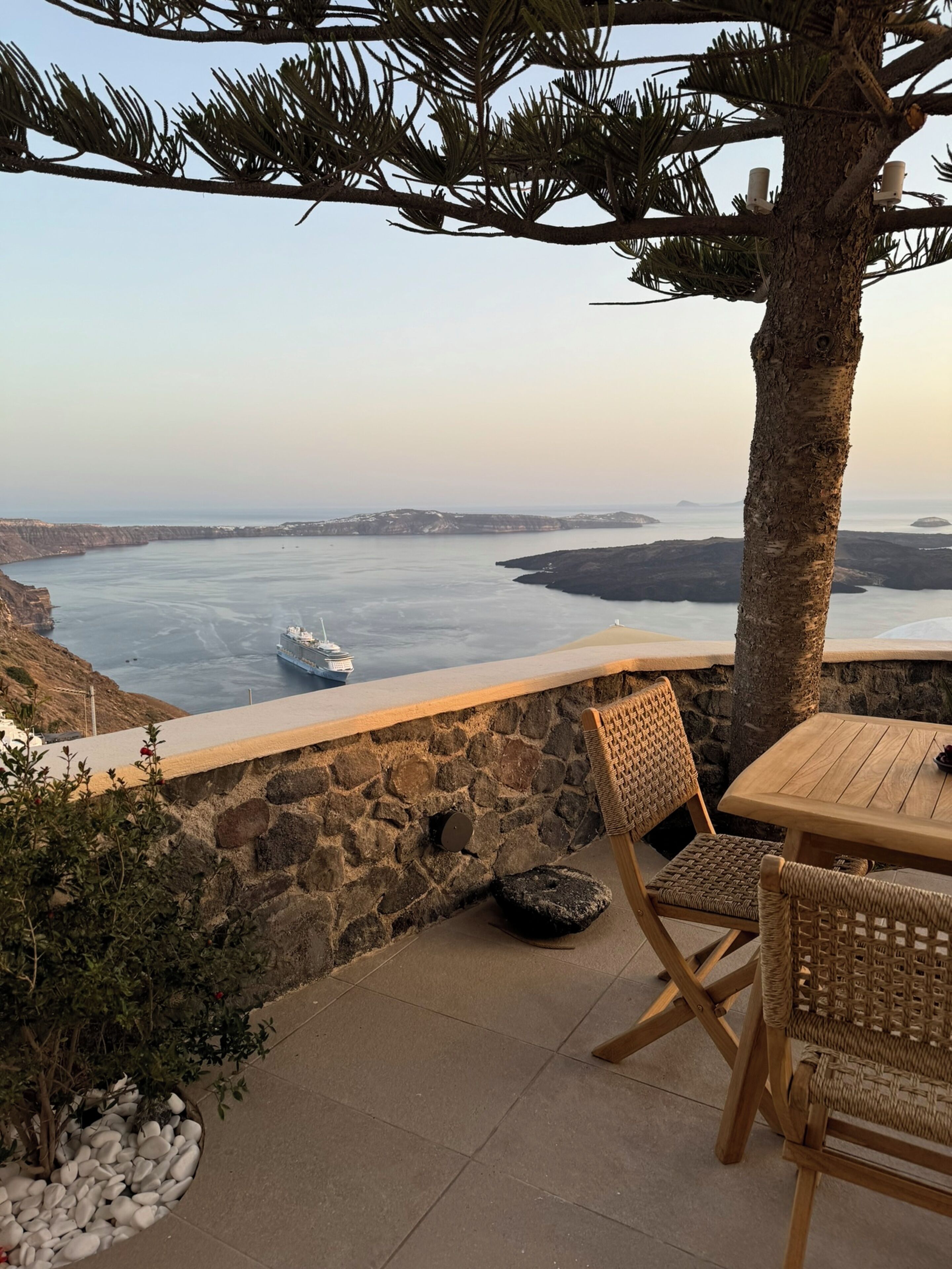 A stone wall with a view of a calm sea and a ship. There is a wooden table and wicker chairs.